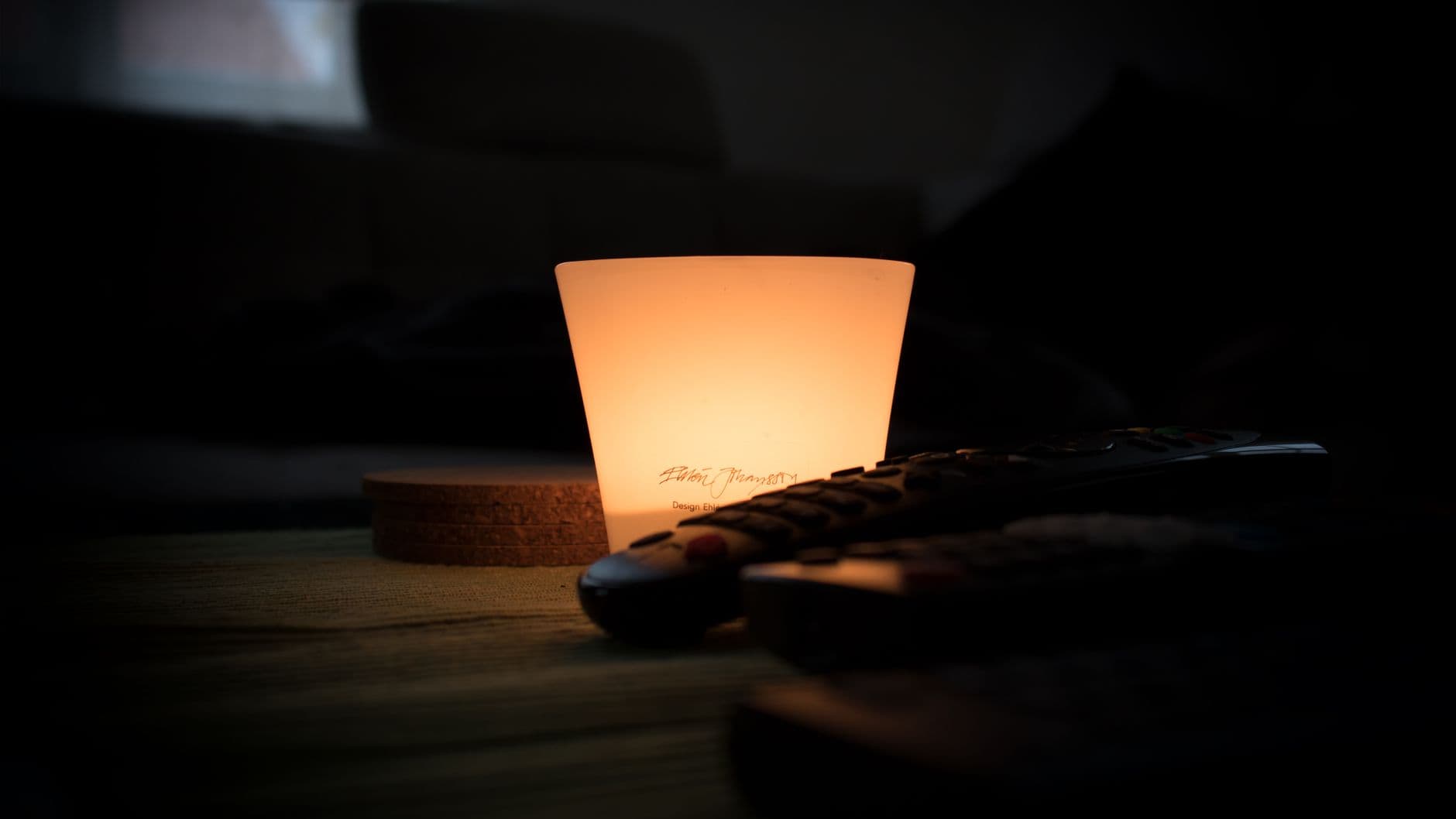 A candle lighting a dark room, with a remote control on the table beside it